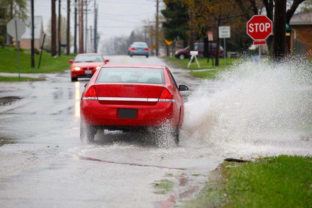 A red car driving through a flooded street, splashing water, with a stop sign in the background.