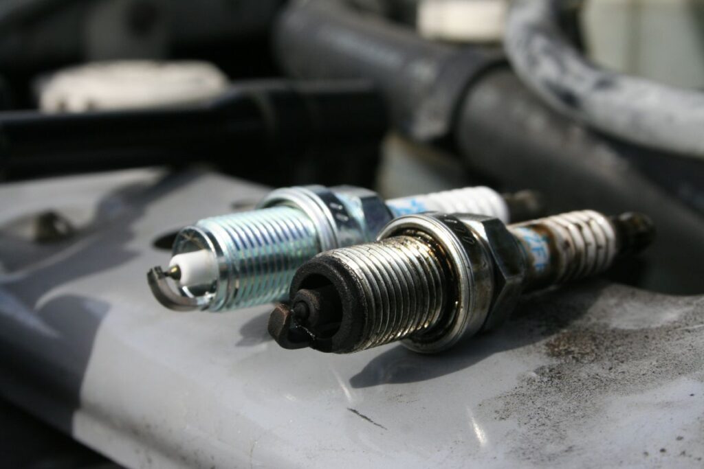 Two spark plugs, one clean and one worn, placed on an engine surface